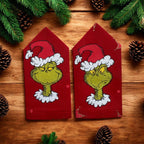 Two red epaulettes with Grinch faces on a wooden surface with pine branches and cones.