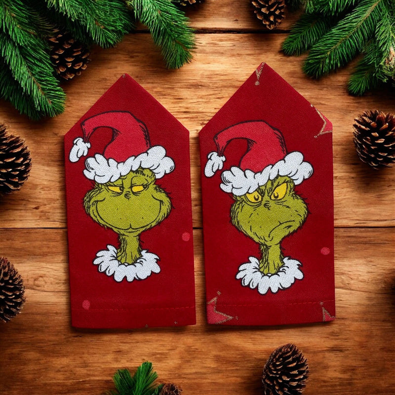 Two red epaulettes with Grinch faces on a wooden surface with pine branches and cones.